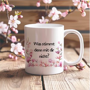 Könnte beinhalten: Weiße Keramiktasse mit floralem Design und dem deutschen Text "Was stimmt denn mit dir nicht?" Die Tasse hat einen Henkel und ist mit rosa Blüten verziert. Die Tasse steht auf einer Holzoberfläche.