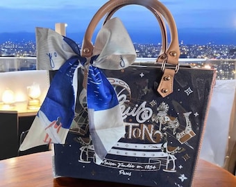 May include: A dark blue structured tote bag with tan leather handles and a large white and blue silk scarf tied to one handle. The bag features a graphic print of a carousel, the text Louis Vuitton, and the phrase Maison Fondée en 1854 Paris.