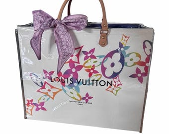 May include: A white Louis Vuitton tote bag featuring a colorful, multi-hued monogram print. The bag has tan leather handles and a light purple patterned ribbon tied to one handle. The text reads LOUIS VUITTON MAISON FONDEE EN 1854 PARIS.