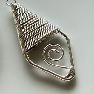 May include: A silver-colored metal pendant featuring a triangular top section tightly wrapped with thin wire and a small circular loop at the apex. The lower portion consists of a bent wire frame containing a centered, free-floating spiral element.