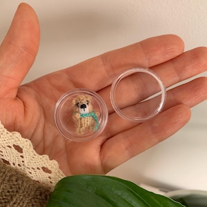 May include: A miniature beige knitted bear figurine with a small teal scarf, enclosed within a clear circular plastic container with a separate matching lid, showcasing intricate yarn texture and a simple black stitched nose and eye details.