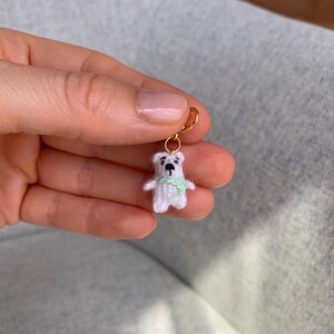 Puede incluir: Un pequeño oso blanco de ganchillo con una bufanda verde claro, sujeto a un gancho de pendiente dorado. El oso tiene ojos negros y una pequeña nariz negra. El pendiente está sujeto por una mano.