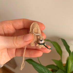 Könnte beinhalten: Handgefertigter, beigefarbener Strickohrring mit einem grünen Schal und schwarzer Nase. Der Ohrring ist an einer beigefarbenen Strickrückseite mit einem silberfarbenen Ohrhaken befestigt. Der Bär ist klein und detailliert.