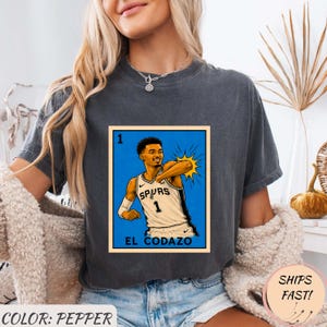 May include: Pepper-colored t-shirt featuring a graphic of a basketball player in a Spurs jersey. The graphic is a stylized illustration with the text "EL CODAZO" at the bottom. The shirt also has the text "SHIPS FAST!"