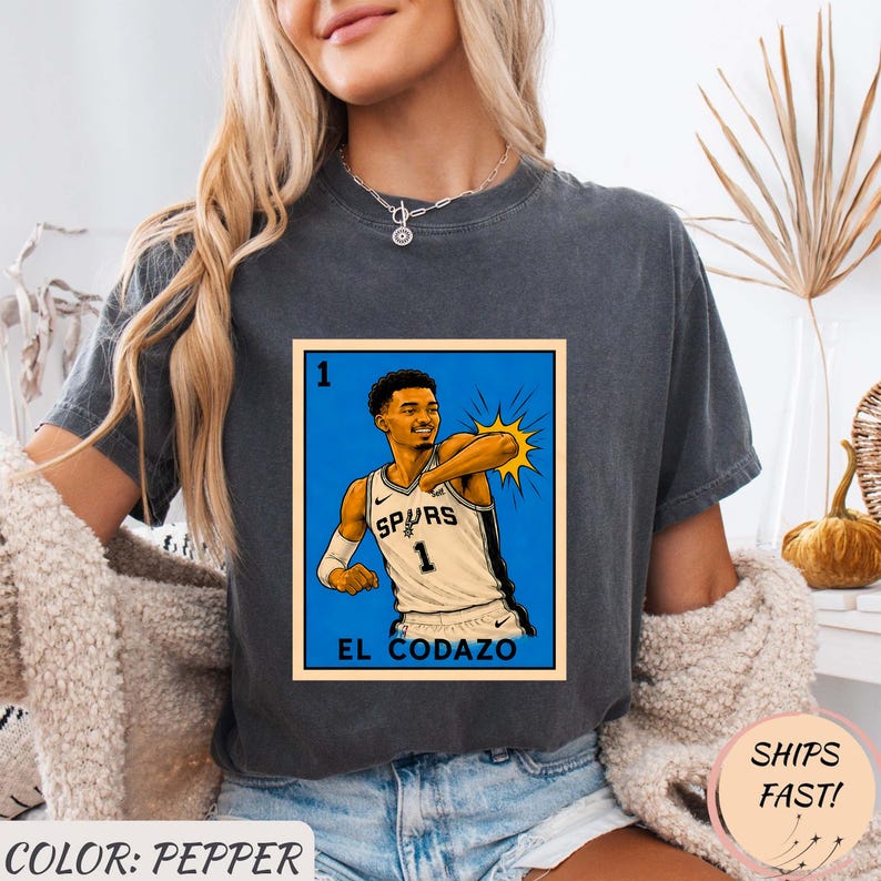 May include: Pepper-colored t-shirt featuring a graphic of a basketball player in a Spurs jersey. The graphic is a stylized illustration with the text "EL CODAZO" at the bottom. The shirt also has the text "SHIPS FAST!"