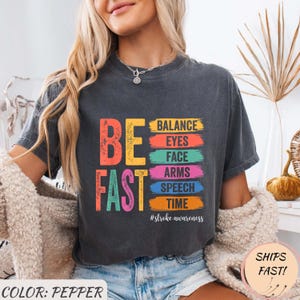 Peut inclure: T-shirt gris foncé avec les mots "BE FAST" en lettres colorées. En dessous, le t-shirt liste "BALANCE, EYES, FACE, ARMS, SPEECH, TIME" dans un arc-en-ciel de couleurs. La phrase "#stroke awareness" est en dessous.