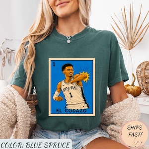 May include: Blue spruce t-shirt featuring a graphic of a basketball player in a Spurs jersey. The graphic is in a style reminiscent of a lottery card, with the text "EL CODAZO" below the image. The shirt also has the text "COLOR: BLUE SPRUCE".