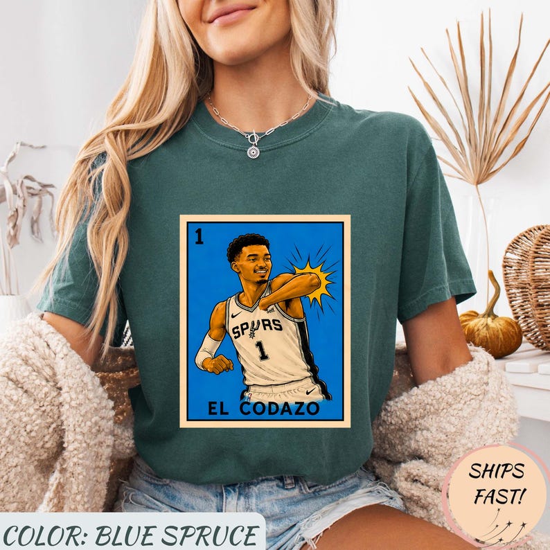 May include: Blue spruce t-shirt featuring a graphic of a basketball player in a Spurs jersey. The graphic is in a style reminiscent of a lottery card, with the text "EL CODAZO" below the image. The shirt also has the text "COLOR: BLUE SPRUCE".