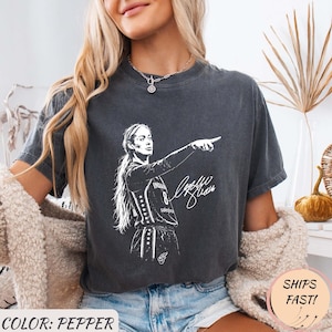 May include: A pepper-colored short-sleeve t-shirt featuring a white graphic illustration of a basketball player with a ponytail wearing a jersey with the number 8, pointing to the side, accompanied by a white signature script design on the front.