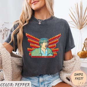 Può includere: T-shirt color pepe con una grafica di una persona con giacca verde e camicia blu, con un design ad ali rosse e arancioni. La maglietta ha il testo "COLOR: PEPPER" in basso.