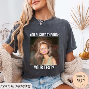 May include: Pepper-colored t-shirt featuring a meme of a doll with messy hair and a displeased expression. The text on the shirt reads "YOU RUSHED THROUGH YOUR TEST?"