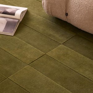 Peut inclure: Tapis rectangulaire vert olive avec un motif géométrique. Un coussin beige texturé est partiellement visible. Un magazine ouvert repose sur le tapis. Dimensions en centimètres.
