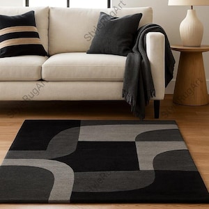 May include: A modern area rug with a geometric design in shades of black, gray, and charcoal. The rug is placed in front of a cream-colored sofa with black and gray throw pillows and a dark gray blanket.