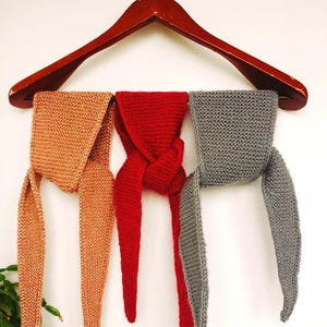 Puede incluir: Tres bufandas de punto en tonos melocotón, rojo y gris cuelgan de una percha de madera. Las bufandas se exhiben contra una pared blanca, con las bufandas melocotón y gris a cada lado de la bufanda roja. Las bufandas tienen extremos largos y puntiagudos.