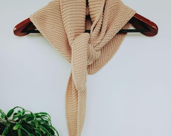 May include: A beige knitted scarf is displayed on a wooden hanger against a white wall. The scarf is folded and knotted, with a textured pattern. The scarf's neutral color complements the natural wood of the hanger. A green plant is visible in the lower left corner.
