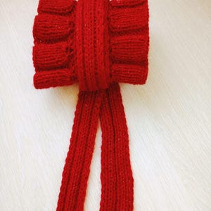 May include: A vibrant red knitted bow, hand-crafted with a textured pattern. The bow has a central knot and two long, rectangular streamers. The yarn has a subtle shimmer, adding a touch of elegance to the accessory.