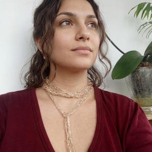 Może przedstawiać: Złoty naszyjnik z wieloma pasmami o długim, drapowanym wzorze. Naszyjnik jest noszony na bordowym topie. Fioletowa orchidea i zielona roślina są widoczne w tle.