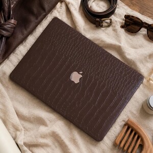 K&ouml;nnte beinhalten: Eine dunkelbraune Laptop-H&uuml;lle mit strukturierter Krokodilleder-Optik auf einem geschlossenen Laptop mit silbernem Apple-Logo. Die Folie bedeckt die gesamte Oberseite des Ger&auml;ts und verleiht ihm eine haptische Oberfl&auml;che mit reptilienartigem Muster.