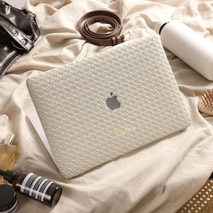May include: A laptop with a beige woven-texture skin featuring a centred Apple logo and the gold-coloured text "Dabrg" printed near the bottom. The laptop is surrounded by a dark brown leather bag, a brown belt, a white bottle, and a hairbrush.