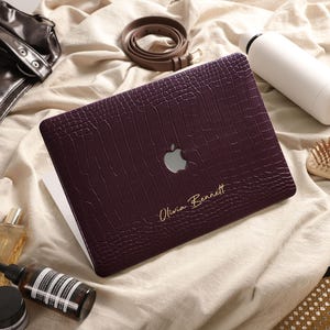 Peut inclure: Une coque d'ordinateur portable violet foncé avec un motif texturé en peau de crocodile, présentant une découpe pour le logo Apple et le nom Olivia Bennett imprimé en doré dans une écriture cursive sur le coin inférieur droit de la surface.