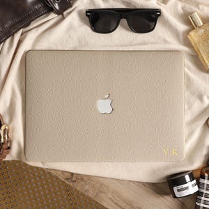 May include: A beige textured laptop skin applied to a laptop with a silver Apple logo in the center. The skin features a pebbled leather-like finish and the gold-colored initials Y.K. are embossed in the bottom right corner of the cover.