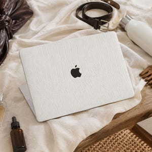 May include: A white laptop skin with a textured, embossed pattern applied to the lid of a closed laptop. The skin features a matte finish and covers the surface while leaving the black Apple logo visible in the centre of the device lid.