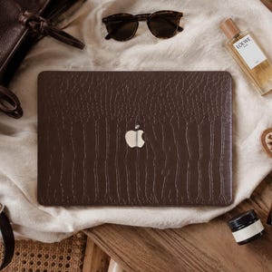 K&ouml;nnte beinhalten: Eine dunkelbraune Laptop-H&uuml;lle mit texturiertem Krokodilmuster und einem mittigen Ausschnitt f&uuml;r das silberne Apple-Logo. Die rechteckige H&uuml;lle ist f&uuml;r den Laptop-Deckel konzipiert und zeigt eine detaillierte, gepr&auml;gte Schuppenoberfl&auml;che.