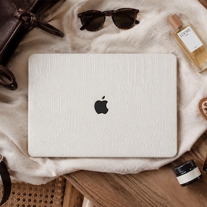 May include: A white laptop skin with a textured crocodile pattern applied to the lid of a closed laptop featuring a black Apple logo in the centre. The laptop is surrounded by a dark brown leather bag, sunglasses, a perfume bottle, and a hairbrush.
