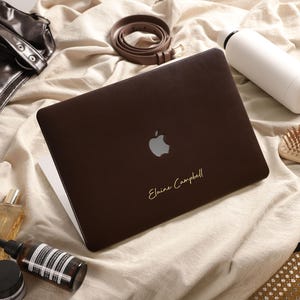 Peut inclure: Un autocollant pour ordinateur portable marron foncé avec un logo Apple argenté et le nom Elaine Campbell imprimé en écriture dorée. L'autocollant est appliqué sur un ordinateur portable entouré d'objets personnels comme une bouteille et une brosse.