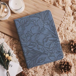May include: A blue leather journal with an embossed floral design. The journal is lying on a fluffy, beige surface, next to a wooden cutting board with a candle and a glass bottle. A small bouquet of dried flowers and a pinecone are also present.