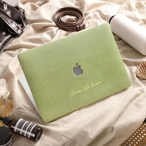 Pode incluir: Uma capa para portátil verde-clara com um padrão texturizado de pele de lagarto e um recorte central para o logótipo da Apple. A capa apresenta texto dourado em cursivo impresso no lado inferior direito, protegendo a superfície exterior.