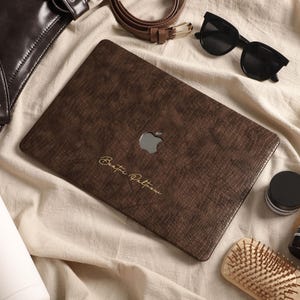 May include: A dark brown textured laptop skin featuring a subtle cross-hatch pattern and the gold cursive text Beata Paltrow centered below the silver Apple logo. The skin is applied to the lid of a laptop, showcasing a sophisticated aesthetic.