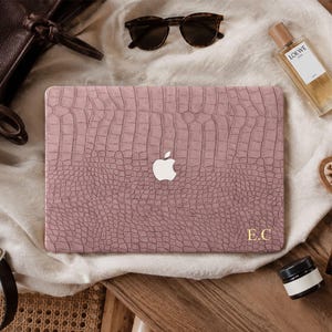 May include: A dusty rose laptop cover with a textured crocodile skin pattern and a central cutout for the Apple logo. The bottom right corner features the gold-colored initials E.C. printed in a serif font on the matte surface of the cover.