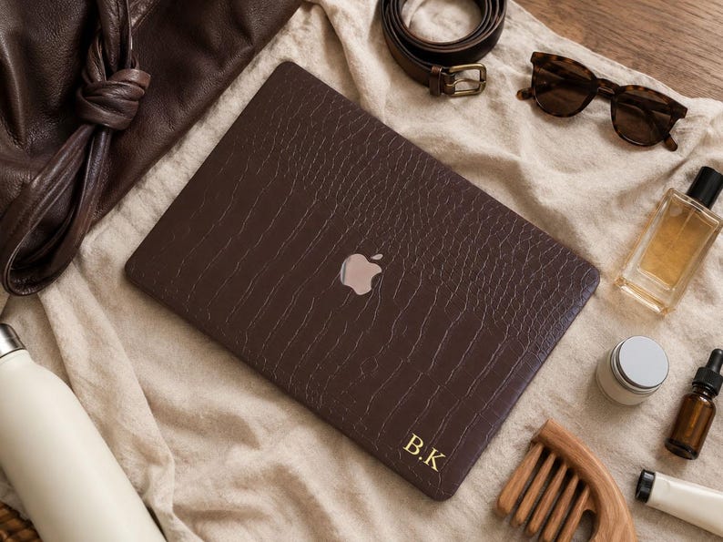 K&ouml;nnte beinhalten: Eine dunkelbraune Laptop-H&uuml;lle mit texturiertem Krokodilleder-Muster, einem silbernen Apple-Logo in der Mitte und den goldenen Initialen B.K in der unteren rechten Ecke, pr&auml;sentiert neben einer Ledertasche, einem G&uuml;rtel und einer Sonnenbrille.