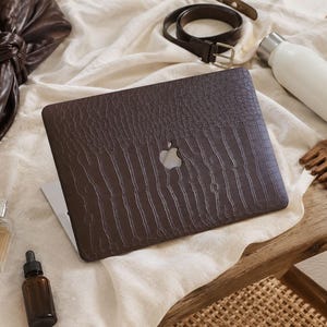 K&ouml;nnte beinhalten: Ein Laptop mit einer dunkelbraunen, strukturierten Krokodilleder-H&uuml;lle auf dem Deckel. Die H&uuml;lle weist ein sich wiederholendes gepr&auml;gtes Schuppenmuster und einen mittigen Ausschnitt auf, der das silberne Apple-Logo auf einer wei&szlig;en Stoffoberfl&auml;che freigibt.