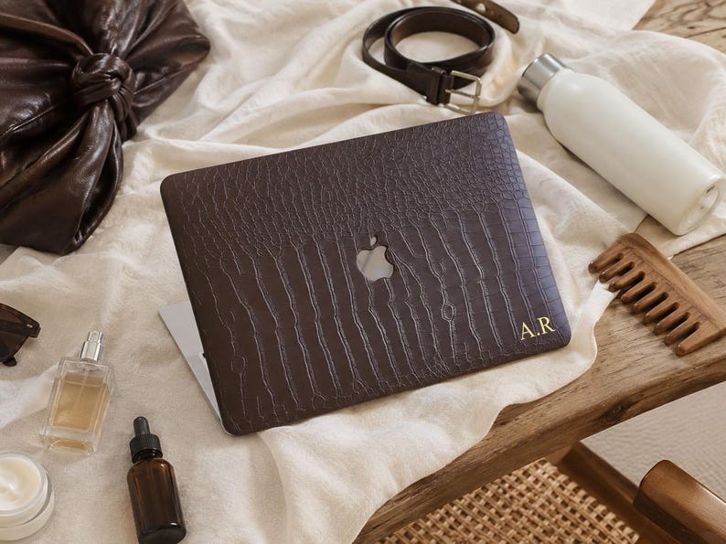 K&ouml;nnte beinhalten: Eine dunkelbraune Laptop-H&uuml;lle mit texturiertem Krokodilleder-Muster und den goldfarbenen Initialen A.R. in der unteren rechten Ecke. Die H&uuml;lle hat einen mittigen Ausschnitt, der das silberne Apple-Logo auf dem Laptop-Deckel freil&auml;sst.