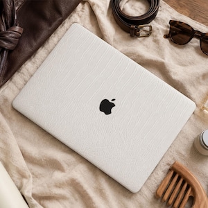 May include: A white laptop skin with a textured, embossed crocodile pattern applied to the lid of a closed laptop. The skin features a precise cutout revealing the black Apple logo in the centre, providing a sleek, protective cover for the device.