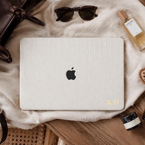 Może przedstawiać: Biała nakładka na laptopa z teksturowanym wzorem skóry krokodyla i czarnym logo Apple na środku. W prawym dolnym rogu znajdują się złote, tłoczone inicjały K.H. Nakładka jest prostokątna i posiada zaokrąglone rogi oraz gładkie wykończenie.
