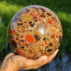 Könnte beinhalten: Eine polierte Steinkugel mit Konglomeratstruktur, in die Fragmente aus roten, orangefarbenen, braunen und durchscheinend gelben Mineralien in einer sandfarbenen Matrix eingebettet sind, was ein Mosaik aus natürlichen Erdtönen ergibt.