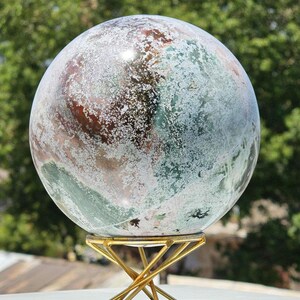 May include: A large decorative sphere with a swirling pattern of green, brown, and white. The sphere is mounted on a gold-coloured metal stand. The stand has a geometric design with a circular top and a crisscross base. The sphere appears to be made of stone.