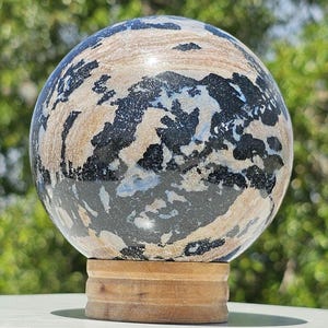 May include: A decorative sphere with a unique pattern of black, tan, and white colors. The sphere is displayed on a small, round, wooden stand. The background is blurred, with green foliage.