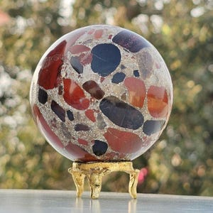 May include: A decorative sphere with a mosaic design of red, black, and orange stones set in a light-colored matrix. The sphere is displayed on a small, ornate gold-colored stand, adding an elegant touch to the piece.