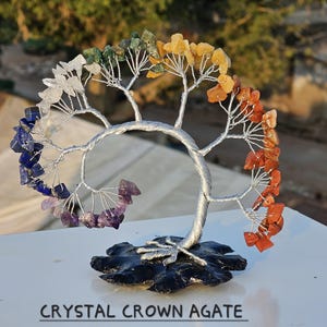 May include: A decorative tree sculpture with a silver wire trunk and branches. The branches are adorned with colorful gemstones in a rainbow arrangement. The base is a black, textured stone. The text "CRYSTAL CROWN AGATE" is at the bottom.