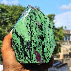 May include: A polished zoisite stone with a pointed top, featuring a vibrant green base colour with prominent black hornblende inclusions and scattered deep magenta ruby patches throughout the surface, displaying a smooth and glossy texture.