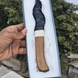 May include: A black obsidian knife with a wooden handle, presented in a grey box with white padding. The handle is wrapped with a white cord. The knife has a curved shape.