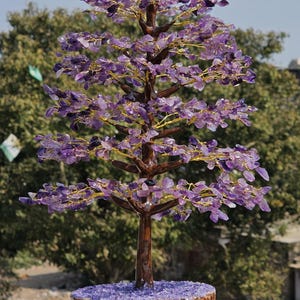 May include: A decorative tree sculpture with a brown trunk and branches, adorned with purple amethyst leaves. The tree sits in a wooden base filled with amethyst chips. The branches are wired with gold-colored wire.