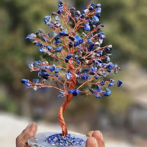 May include: A handcrafted wire tree sculpture with a copper-colored trunk and branches. The tree's leaves are made of blue lapis lazuli stones. The tree is mounted on a white and gray agate slice base.