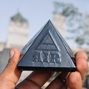 May include: A black pyramid with the word "AIR" and a triangle symbol engraved on its surface. The pyramid is held in a hand against a blurred outdoor background.