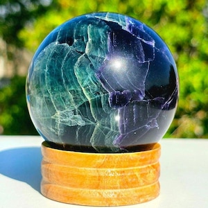 May include: A polished fluorite sphere, showcasing a blend of blue, green, and purple hues. The crystal ball sits on a layered wooden stand, creating a striking contrast. The sphere's surface displays natural patterns and textures.