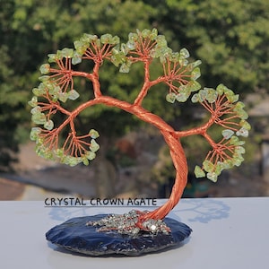 Puede incluir: Una escultura de árbol de alambre hecha a mano con un tronco y ramas de color cobre, adornada con hojas de piedras preciosas verdes. El árbol se asienta sobre una base de piedra azul oscuro. El texto "CRYSTAL CROWN AGATE" es visible.